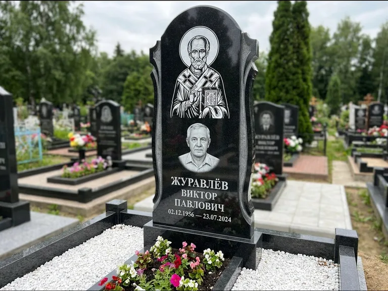 Памятник с гравировкой иконы: стела с образом в полный рост на обороте — гравировка образа Матроны Московской, низкие кусты спиреи в цвету, работа «Лебедева» в Самаре